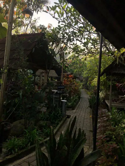 Jalan setapak paving di antara tanaman tropis, dengan atap bangunan di sisi kanan