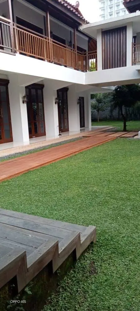 Halaman rumput dengan dek kayu dan jalan bata merah di depan bangunan dua lantai