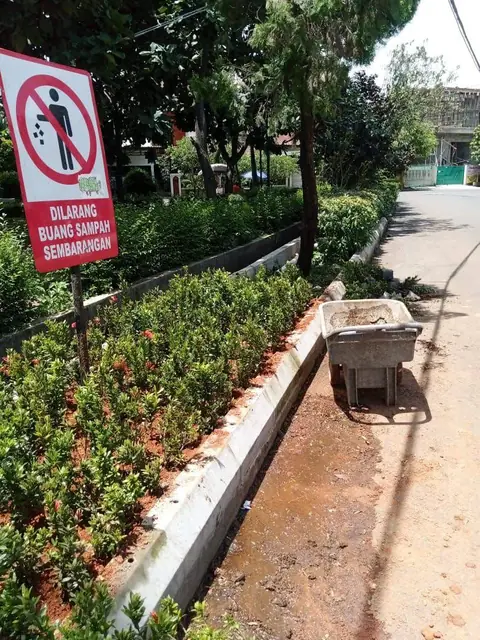 Tanaman pagar yang baru ditanam di samping trotoar, ada gerobak dorong dan papan larangan buang sampah