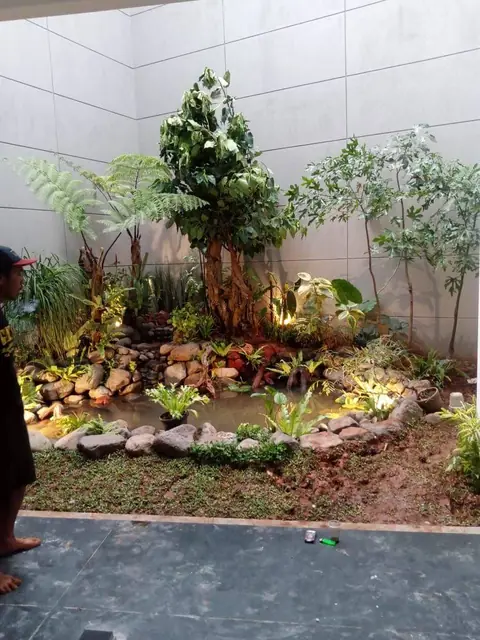 Kolam kecil dikelilingi batu, tanaman, dan pohon di sudut dekat dinding abu-abu